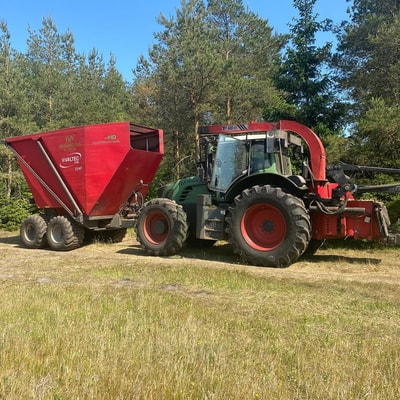 Traktor med flishugger og rød flisvogn parkeret ved nåleskov