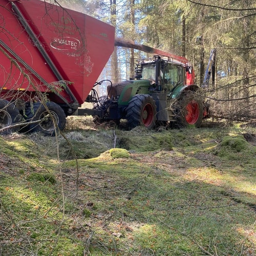 Traktor med flishugger blæser træflis op i rød vogn under skovarbejde