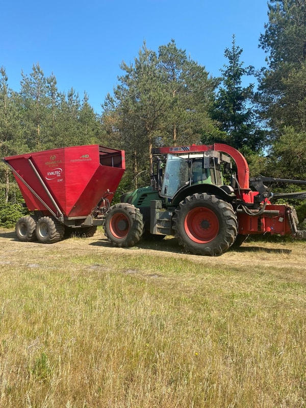 Traktor med flishugger og rød flisvogn parkeret ved nåleskov